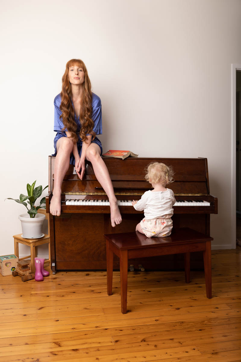 Emma sitting on top of an upright piano with her feet on the keys, while her toddler kneels on the stool below, reaching for the keyboard.