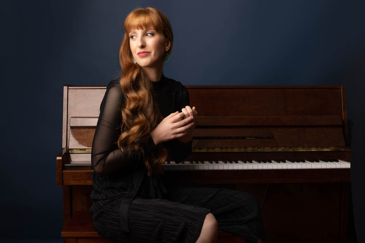 Emma seated at an upright piano in a navy-toned studio.